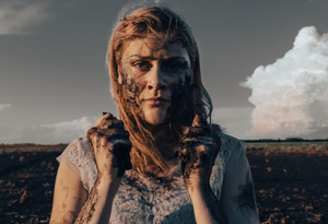 A woman stands in a field, wearing a blue lace dress. She has dirt smeared on her face and hands.