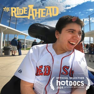 A young man with brown hair smiling in his wheelchair wearing a Red Sox jersey at a baseball game.