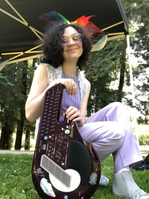 Alex kneels in the grass with a guitar. They are wearing a lilac outfit and heart framed glasses.