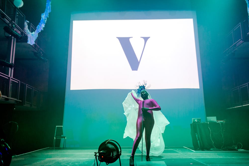 Ebony is wearing a hot pink body suit on a lit up stage with a giant "V" projected on the background.