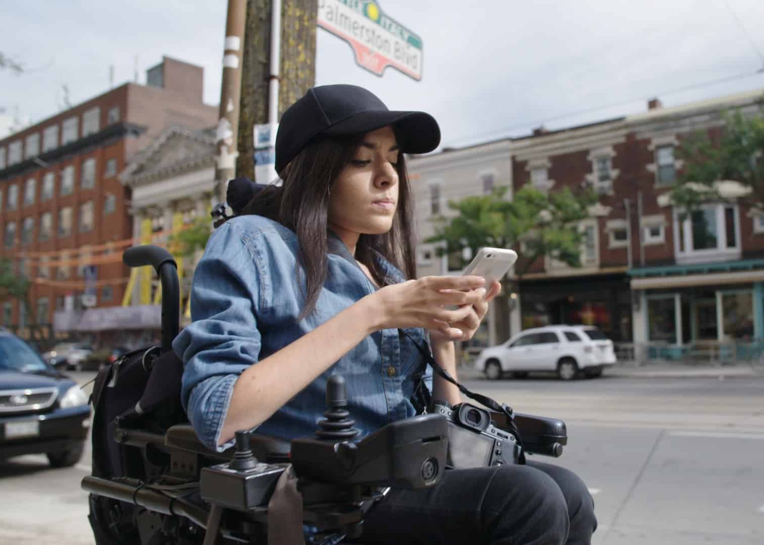 Maayan Ziv sitting in a power chair on an urban street and scrolling on her phone.