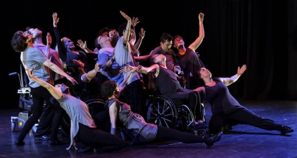 Dancers of all abilities are frozen in a dramatic pose, arms stretched wide, on a stage with blue lighting effects.