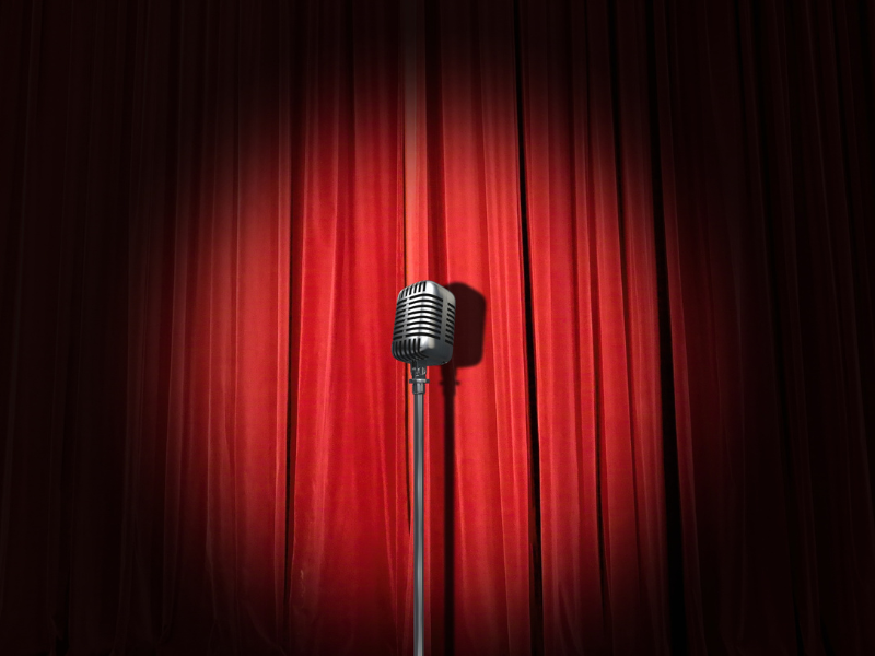 A lone microphone sitting in a spotlight on a stage with red curtains