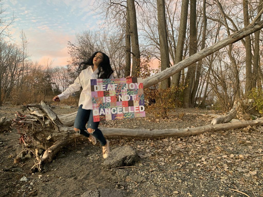 A woman leaping in the air with a sign that reads Deaf Joy is not cancelled