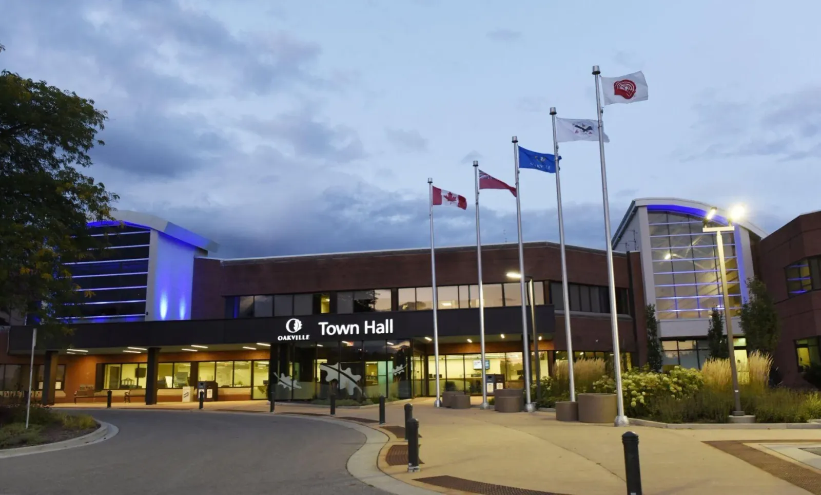 A view of the front of Oakville Town Hall's building. Flag poles are visible and the town's marquee is lit
