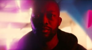 A Black man standing in a pink glowing space and facing the camera. 