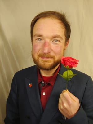 D'arcy is wearing a navy and red suit, holding a plastic rose in his left hand.