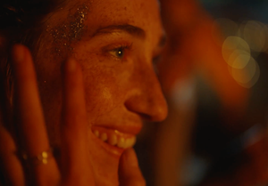 A still from the short film "Better." A woman smiles, her sister applying glitter to her face.